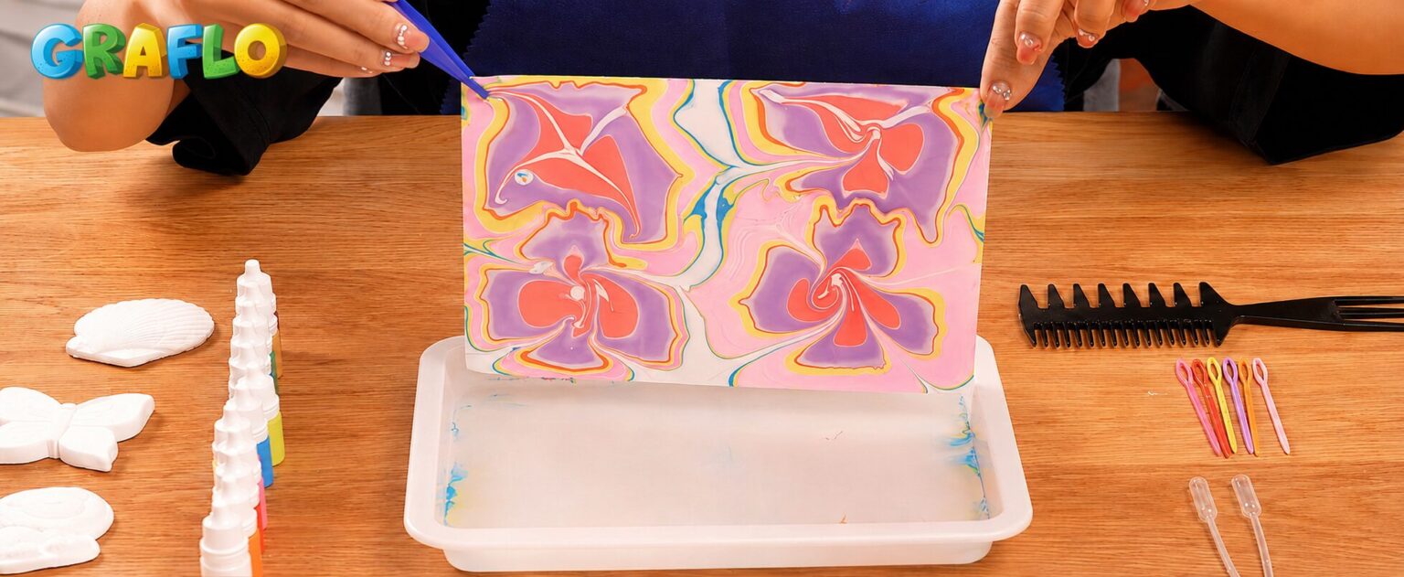 Hands lifting a finished marbled print from the GRAFLO tray, with the comb, awls and pipettes on the table